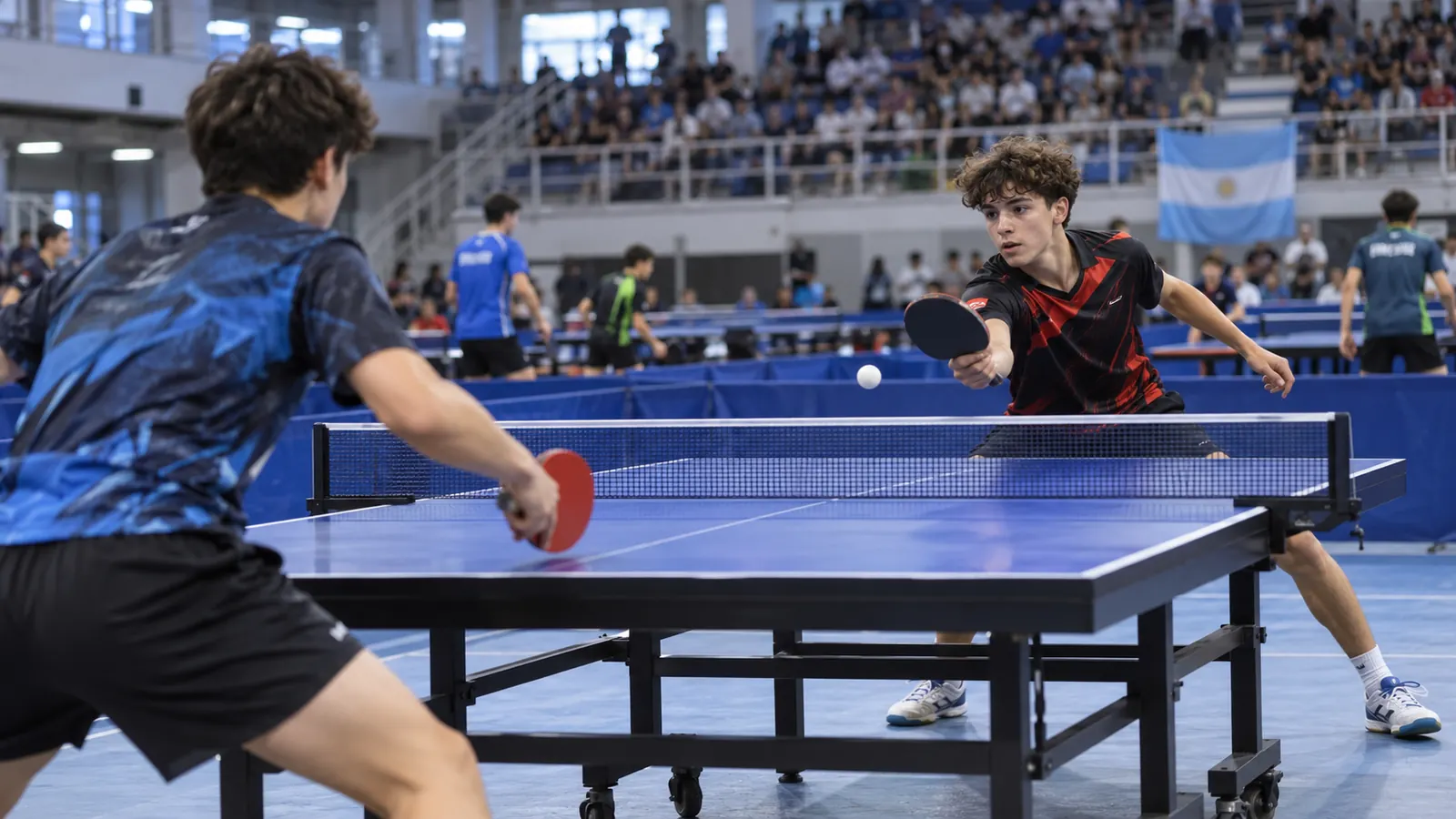 Dos jugadores juveniles disputan un punto de tenis de mesa en un polideportivo con público en las tribunas